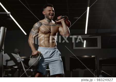 Portrait of young muscular man training shirtless in gym indoors. Hand exercises with kettlebell. Relief body shape. Concept of sport, workout, strength Portrait of young muscular man training shirtless in gym indoors. Hand exercises with kettlebell. Relief body shape. Concept of sport, workout, strength 98324927