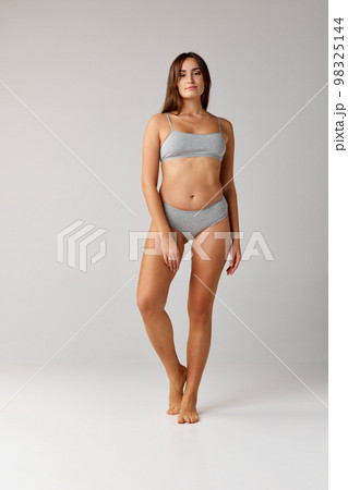 Self-care. Portrait of young beautiful girl with slim body posing in cotton underwear over grey studio background. Concept of body and skin care, fitness Self-care. Portrait of young beautiful girl with slim body posing in cotton underwear over grey studio background. Concept of body and skin care, fitness 98325144