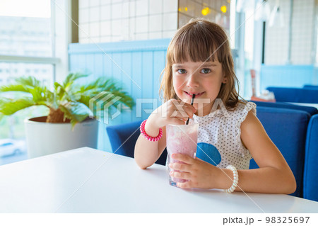 Little girl sitting at a table in a restaurant, with a milkshake in her hands. Healthy nutrition and lifestyle Little girl sitting at a table in a restaurant, with a milkshake in her hands. Healthy nutrition and lifestyle 98325697