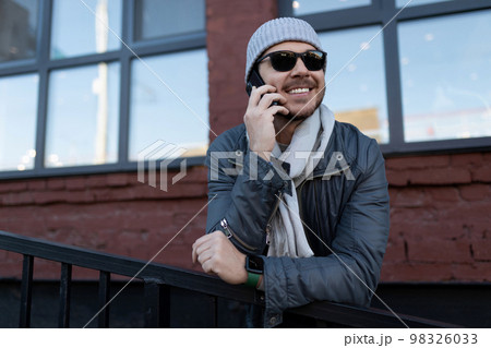 a man in sunglasses and a hat with a scarf speaks with a smile on the phone outside 98326033