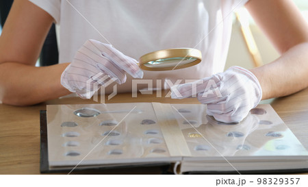 Numismatist in gloves examines collection of coins through magnifying glass. Numismatist in gloves examines collection of coins through magnifying glass. 98329357
