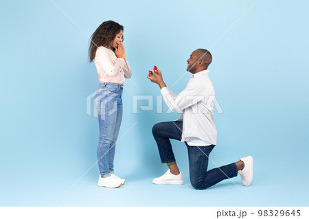 Loving black man standing on one knee and offering engagement ring to his beloved woman on blue background, side view Loving black man standing on one knee and offering engagement ring to his beloved woman on blue background, side view 98329645