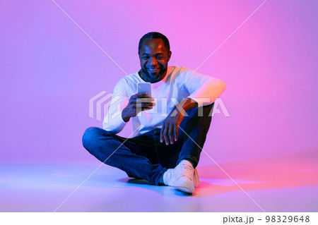 Happy black man sitting on floor, using smartphone and smiling 98329648