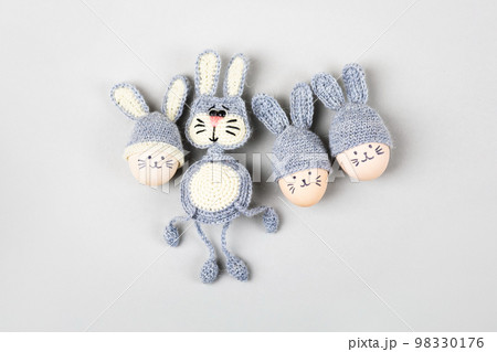 Three Easter eggs with funny faces and gray crocheted hats with bunny ears and a crocheted gray bunny on a gray background. Happy Easter concept. Greeting card. 98330176