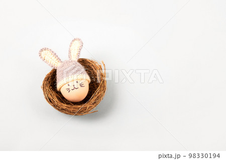 Easter egg with funny face and gray crocheted hat with bunny ears in a decorative nest of sisal on a gray background. Happy Easter concept. Greeting card. 98330194
