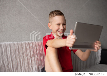 A cute boy in a red T-shirt is sitting playing on a tablet and laughing 98330435