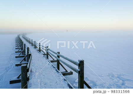 Wooden boardwalk on the frozen lake leading to the bird watching tower, winter sunset landscape Wooden boardwalk on the frozen lake leading to the bird watching tower, winter sunset landscape 98331566