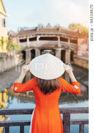 Woman traveler wearing Ao Dai Vietnamese dress sightseeing at Japanese covered bridge in Hoi An town, Vietnam. landmark and popular for tourist attractions. Vietnam and Southeast Asia travel concept 98338670