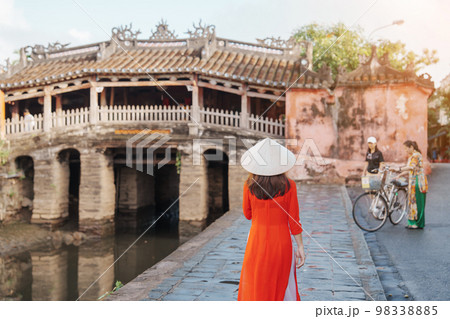 Woman traveler wearing Ao Dai Vietnamese dress sightseeing at Japanese covered bridge in Hoi An town, Vietnam. landmark and popular for tourist attractions. Vietnam and Southeast Asia travel concept 98338885