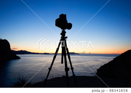 Photo camera silhouette on tripod at rocky beach 98341658