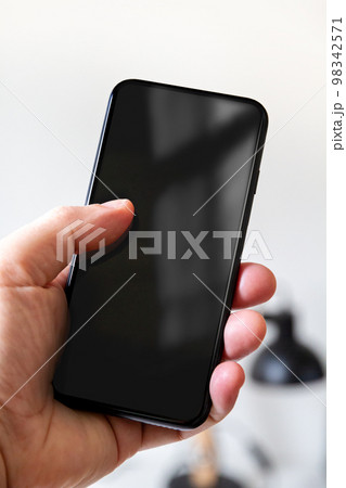 Hand holding a smartphone with blank black screen. Office background. 98342571