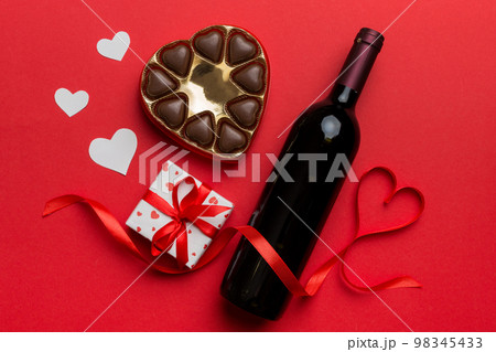 Bottle of red wine on colored background for Valentine Day with gift and chocolate. Heart shaped with gift box of chocolates top view with copy space Bottle of red wine on colored background for Valentine Day with gift and chocolate. Heart shaped with gift box of chocolates top view with copy space 98345433