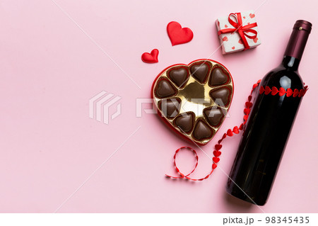 Bottle of red wine on colored background for Valentine Day with gift and chocolate. Heart shaped with gift box of chocolates top view with copy space Bottle of red wine on colored background for Valentine Day with gift and chocolate. Heart shaped with gift box of chocolates top view with copy space 98345435