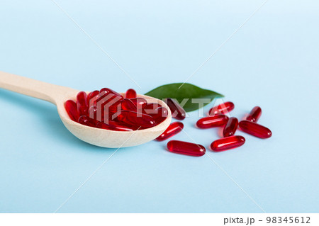 Vitamin capsules in a spoon on a colored background. Pills served as a healthy meal. Red soft gel vitamin supplement capsules on spoon 98345612