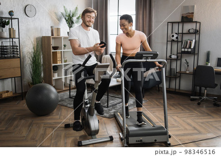 Happy smiling couple doing cardio on stationary bike and treadmill using smart phone 98346516