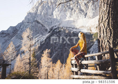 Woman hiker looking away from the camera at the view of the mountains, sitting on a wooden fence enjoying the sun 98351506