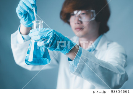 Young asian male scient hand hold erlenmeyer flask with stirring rod filled with blue sample chemicals in chemistry science laboratory. Glassware in medical research. Scientific lab for Biotechnology. 98354661