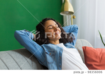 Beautiful hispanic woman at home close up with closed eyes relaxing sitting on couch, woman with hands behind head smiling in living room. Beautiful hispanic woman at home close up with closed eyes relaxing sitting on couch, woman with hands behind head smiling in living room. 98356006