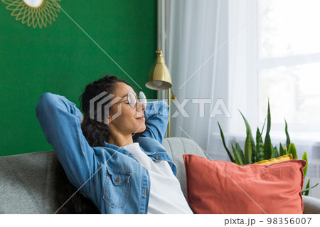 Beautiful hispanic woman at home close up with closed eyes relaxing sitting on couch, woman with hands behind head smiling in living room. Beautiful hispanic woman at home close up with closed eyes relaxing sitting on couch, woman with hands behind head smiling in living room. 98356007