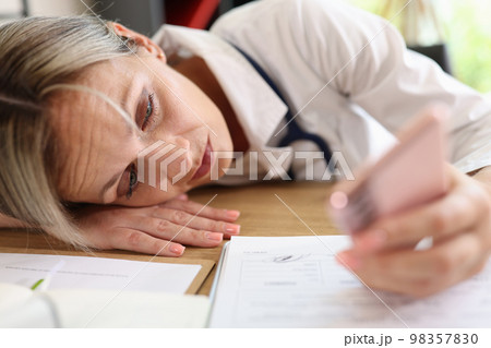 Tired female doctor laying on her work desk and looking at smartphone screen. 98357830