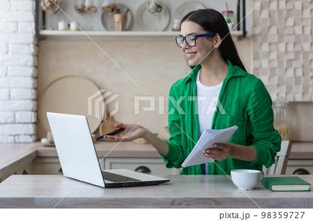 A young female student, a freelancer studies, works online at home from a laptop. He sits at the table, listens to the lecture, holds a notebook with a pen in his hands, writes down, explains. A young female student, a freelancer studies, works online at home from a laptop. He sits at the table, listens to the lecture, holds a notebook with a pen in his hands, writes down, explains. 98359727