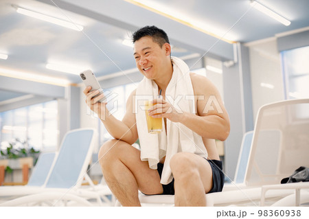 Man has a white towel on his shoulders. Asian man sitting on a sunbed in indoor swimming pool and holding a glass with orange juice. Professionale male swimmer using a phone. Man has a white towel on his shoulders. Asian man sitting on a sunbed in indoor swimming pool and holding a glass with orange juice. Professionale male swimmer using a phone. 98360938