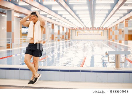 Man wearing special hat and goggles for swimming. Asian man standing in indoor swimming pool with a white towel on his shoulders. Professionale male swimmer is going to swim. 98360943
