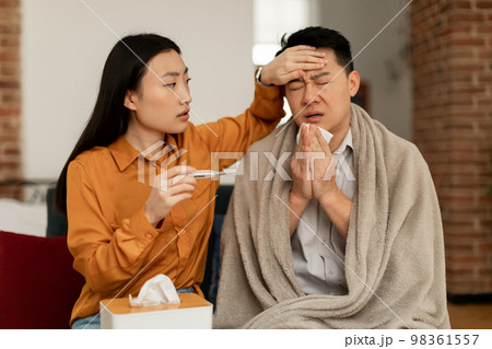 Asian woman treating sick husband, holding thermometer and touching man forehead, sitting on couch at home Asian woman treating sick husband, holding thermometer and touching man forehead, sitting on couch at home 98361557