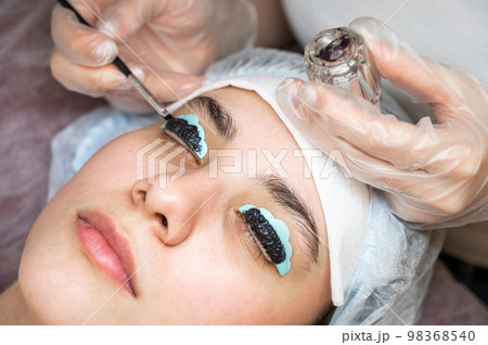 Close-up portrait of a woman on eyelash lamination procedure. The master applies tint to the eyelashes.  98368540