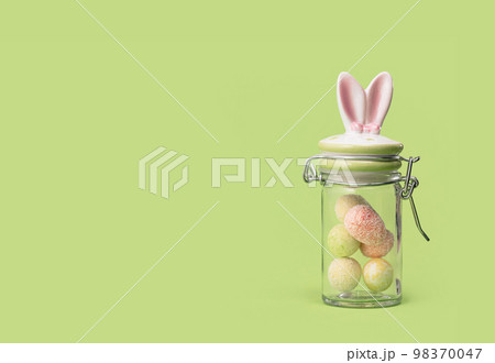 Easter bunny ears sweets glass jar with tiny eggs inside on spring yellow green edgeless background. Happy Easter holiday concept. Easter chocolate treats web wide banner Easter bunny ears sweets glass jar with tiny eggs inside on spring yellow green edgeless background. Happy Easter holiday concept. Easter chocolate treats web wide banner 98370047