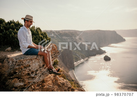 Digital nomad, man in the hat, a businessman with a laptop sits on the rocks by the sea during sunset, makes a business transaction online from a distance. Remote work on vacation. 98370707