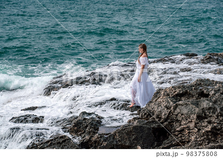 A woman stands on a rock in the sea during a storm. Dressed in a white long dress, the waves break on the rocks and white spray rises. 98375808