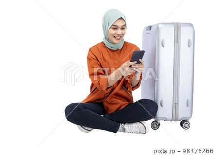 Asian Muslim woman in a headscarf sitting with a suitcase while using a mobile phone 98376266