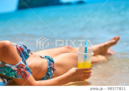 A beautiful slender girl in a swimsuit enjoys a tan while lying on a tropical beach, holding a glass of pineapple juice in her hand 98377821