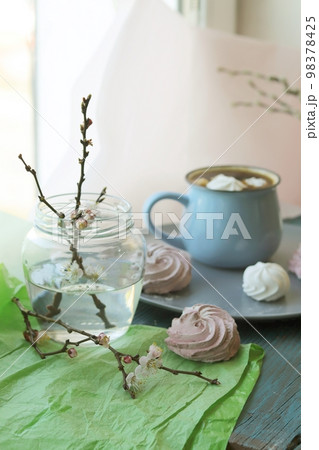 Cup of coffee with meringue, flowering cherry branches in water in a glass jar, sweet dessert on a plate on a wooden windowsill, spring season, home comfort 98378425