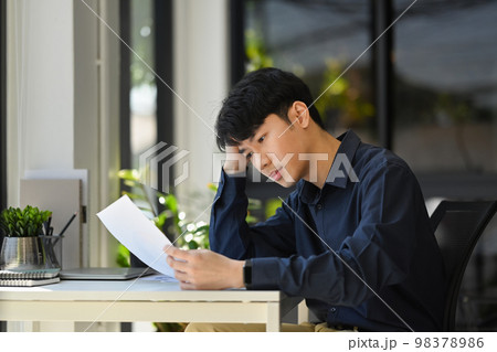 Frustrated asian male office worker feeling stressed, checking financial report at his office desk 98378986