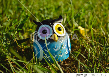 A toy of a surprised blue-colored owl with multi-colored eyes close-up stands in the middle of green grass in the light of the evening sun 98380405