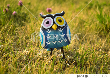 A toy of a surprised blue-colored owl with multi-colored eyes close-up stands in the middle of the grass in the light of the evening sun 98380409
