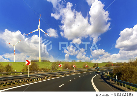 Wide highway with moving car stretching to horizon by green hills with high wind turbines on blue cloudy sky background. Modern technologies, renewable energy source and ecological problems solving. 98382198