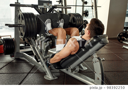 Young man is engaged on a modern simulator in the gym. Male athlete is doing exercises for legs with training machine. 98392250