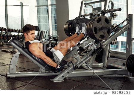 Man Using A Press Machine In A Fitness Club. Strong man doing an exercise on its feet in the simulator. Side view. 98392252