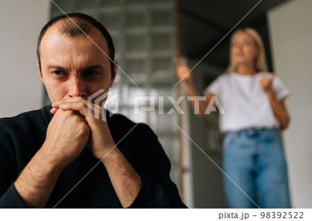 Close-up face of serious depressed husband sitting on bed looking at camera ignoring angry jealous wife arguing blaming upset man of problems at home, standing on blurred background. 98392522