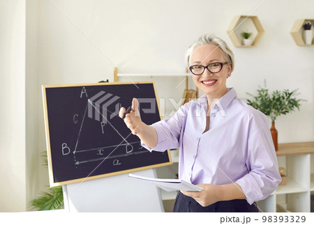 Cheerful gray-haired woman teacher or private tutor stands at blackboard with geometric drawings 98393329