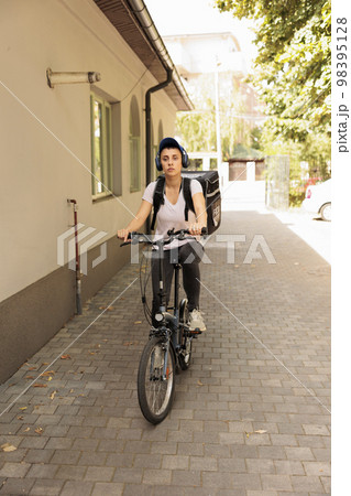 Takeaway food courier in headphones delivering order by bike, front view. Meal delivery service employee riding bicycle with customer order in backpack portrait, woman delivering lunch 98395128