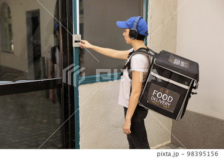 Food delivery service woman courier ringing building doorbell, waiting for client outdoors. Girl in headphones with thermal backpack standing in front of door, delivering takeaway lunch 98395156