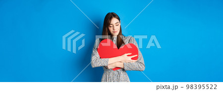 Valentines day. Romantinc girl in dress hugging big red heart cutout, close eyes and smile with dreamy face, imaging sensual date, standing on blue background 98395522
