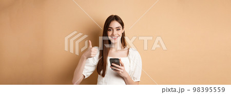 Online shopping concept. Smiling happy woman showing thumb up after using smartphone, holding phone and say yes, standing on beige background Online shopping concept. Smiling happy woman showing thumb up after using smartphone, holding phone and say yes, standing on beige background 98395559