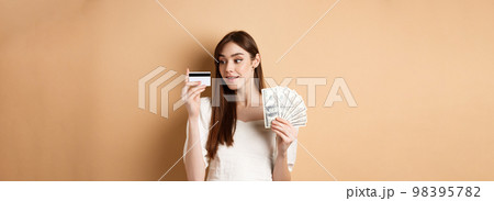 Dreamy girl looking at plastic credit card and thinking of shopping, holding dollar bills, standing on beige background Dreamy girl looking at plastic credit card and thinking of shopping, holding dollar bills, standing on beige background 98395782