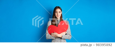 Valentines day. Attractive young woman searching for love, holding big red heart cutout and smiling at camera, standing in dress on blue background 98395892