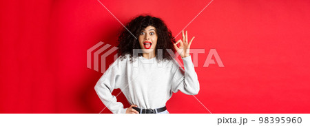 Excited pretty woman say yes, showing okay gesture and looking enthusiastic, agree or like something, making compliment, standing on red background Excited pretty woman say yes, showing okay gesture and looking enthusiastic, agree or like something, making compliment, standing on red background 98395960